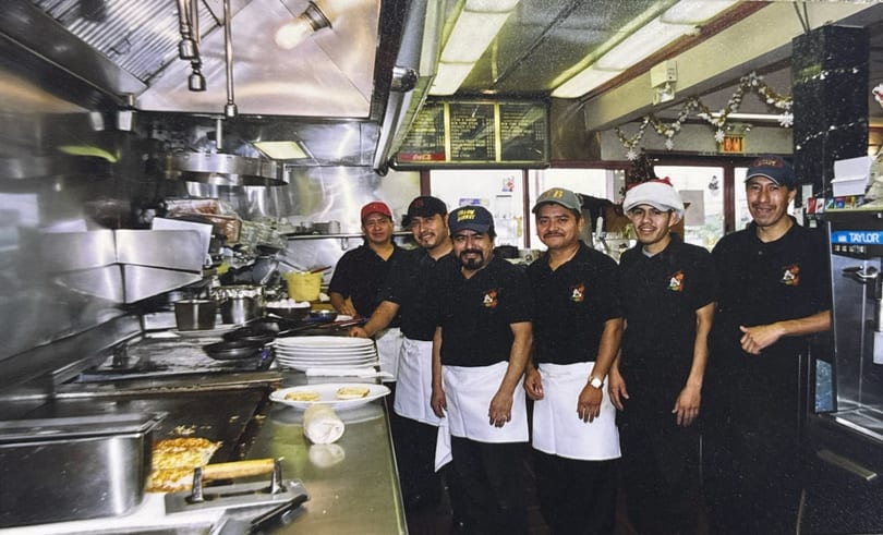 Six male kitchen staff in black shirts and white aprons smiling in a busy restaurant kitchen.