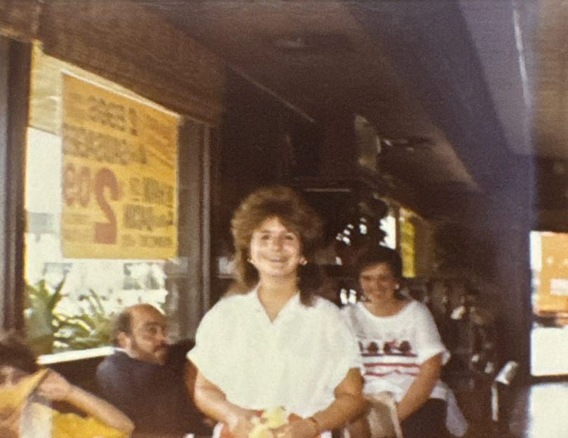 Smiling woman with big hair in a white shirt, with other patrons in a restaurant.