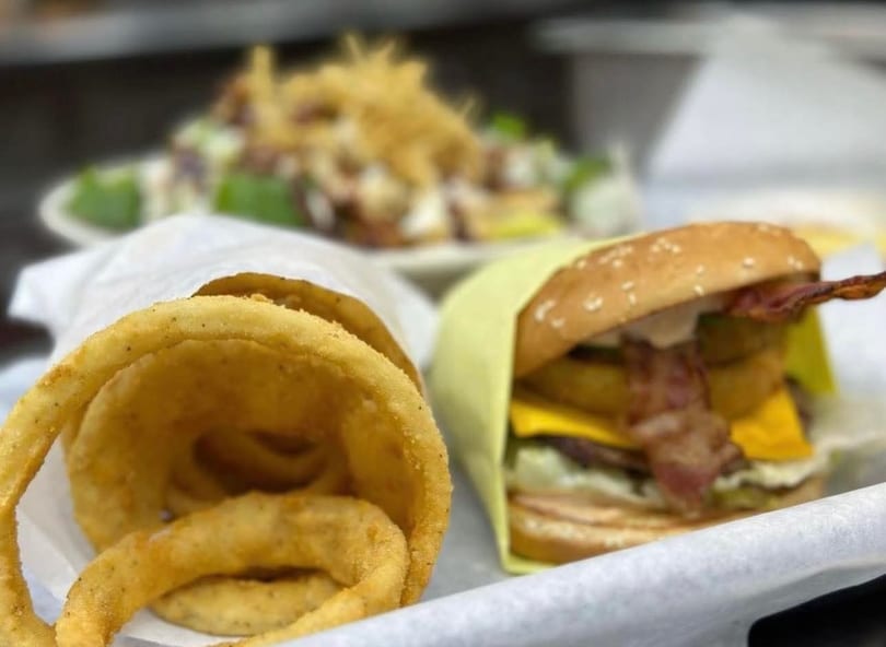 Crispy onion rings in a paper bag next to a bacon cheeseburger with a salad in the background.