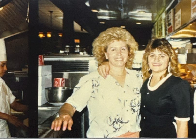 Two women smiling, one with an arm around the other, in a restaurant kitchen with a chef.