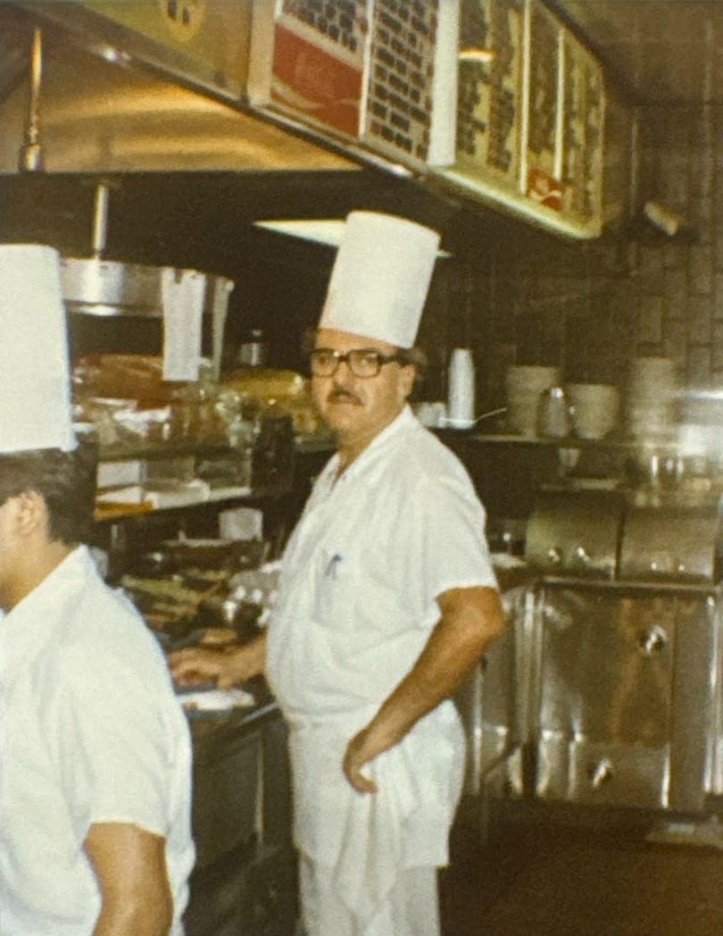 A chef in a tall white hat and glasses stands in a busy restaurant kitchen.
