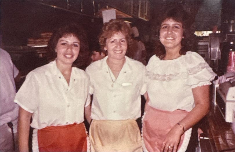 Three smiling women in white shirts and aprons stand together in a restaurant kitchen.