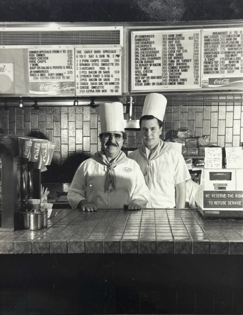 Two chefs in tall white hats and uniforms stand behind a tiled counter with menus above.