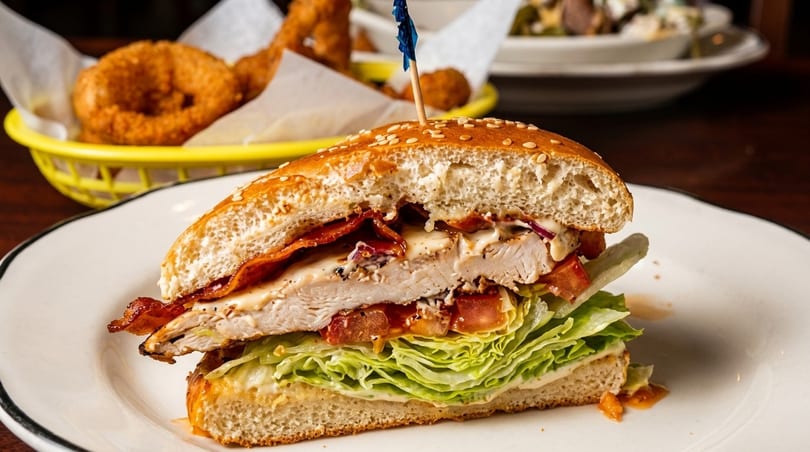 A sliced chicken sandwich with bacon, lettuce, tomato, and sauce on a sesame bun, with onion rings in the background.