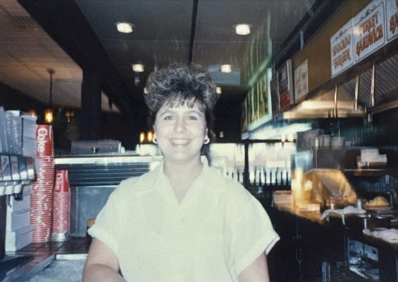 Smiling woman with curly hair in a diner, with soda dispensers and menu boards in the background.