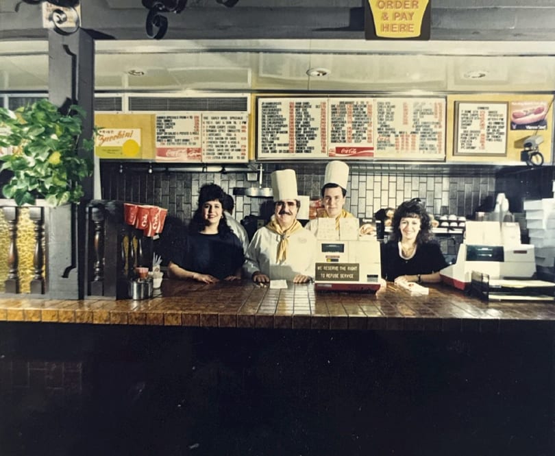 Four smiling restaurant staff members, two in chef hats, stand behind a counter with menus above.