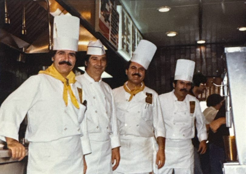 Four chefs in white uniforms and hats, two with yellow scarves, stand smiling in a kitchen.